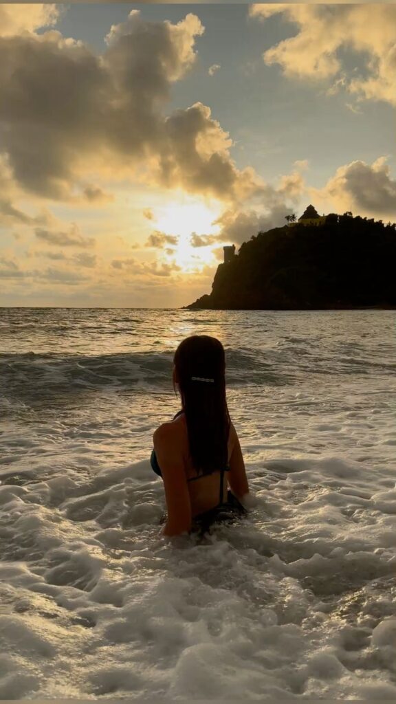 Woman in ocean at sunset, looking towards a hill with buildings silhouetted against a dramatic cloudy sky.