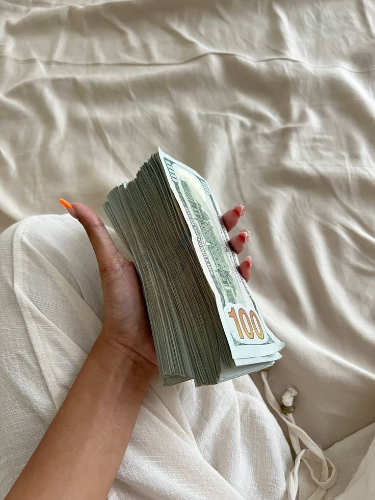 Hand holding a stack of 100-dollar bills on a beige fabric background.