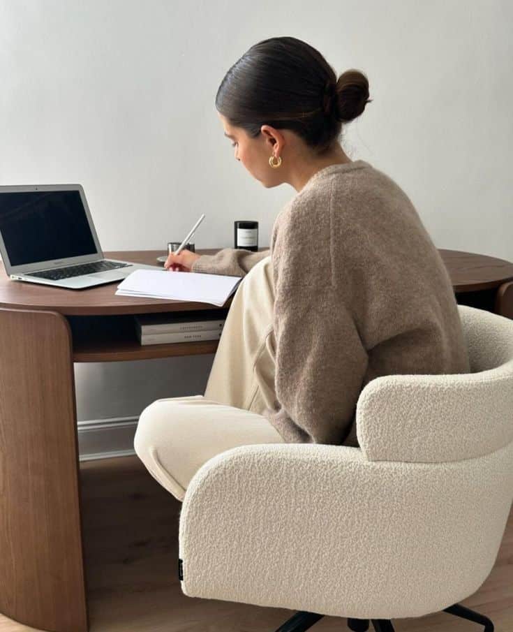 Woman writing at a desk with a laptop, in a cozy chair, in a home office setting.