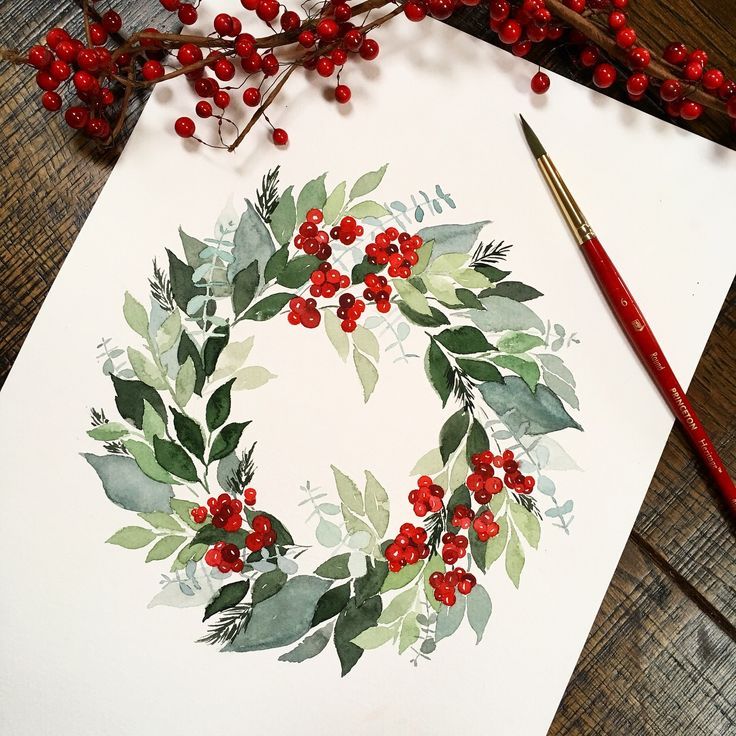 Watercolor holly wreath with red berries on paper beside paintbrush, festive holiday art.
