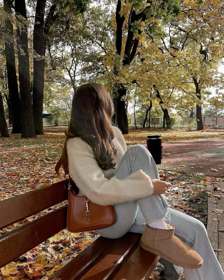 Woman sitting on park bench in autumn, wearing cozy sweater and boots, surrounded by colorful fallen leaves.