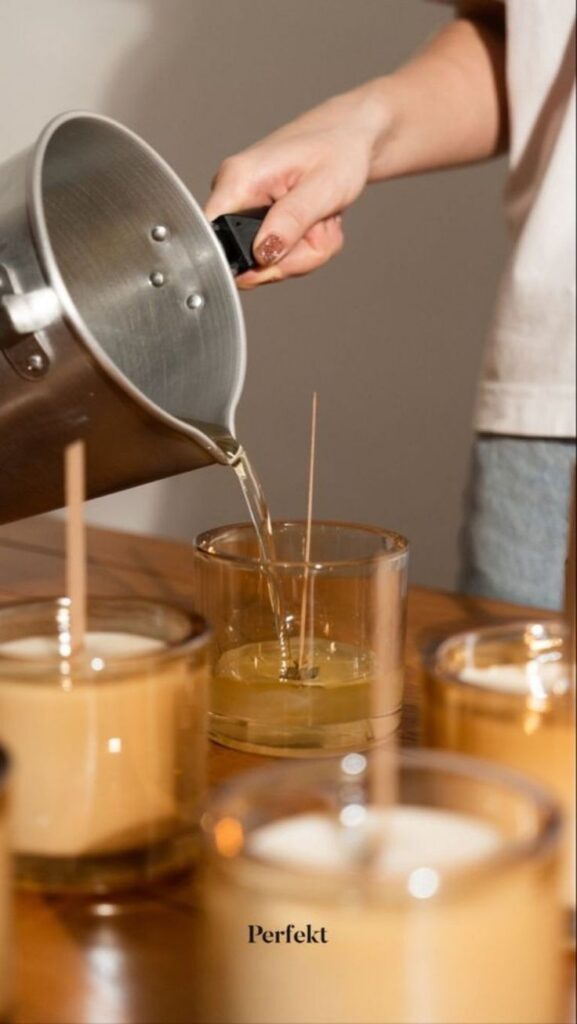 Person pouring wax into glass jars for candle making on a wooden table.