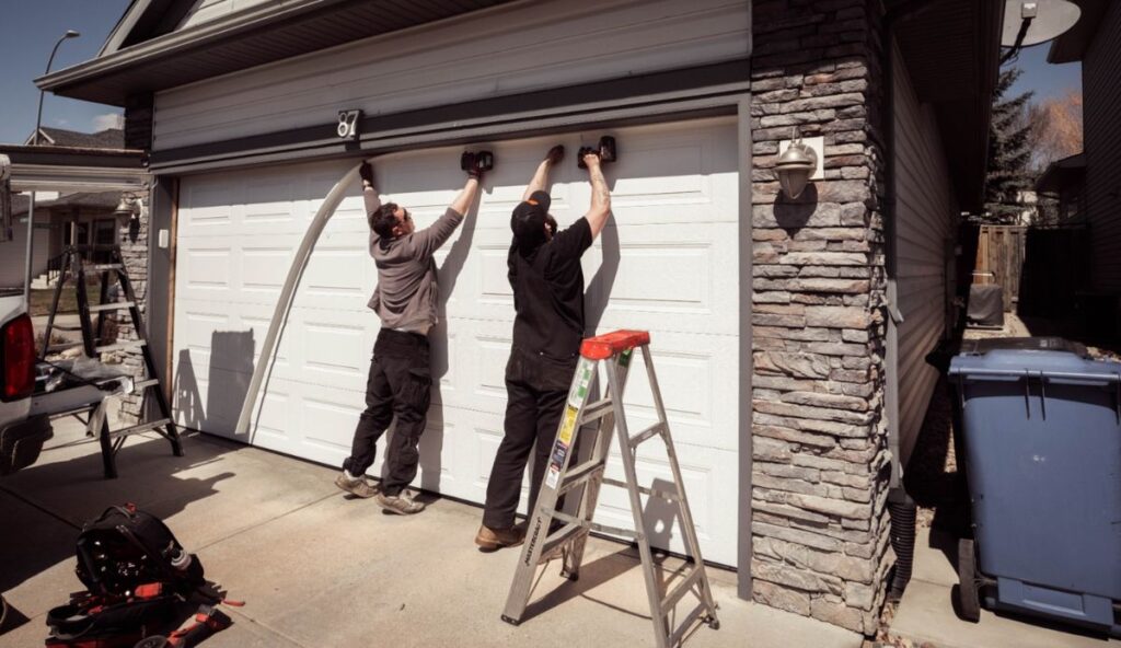 Two technicians installing a white garage door with tools and ladder nearby.