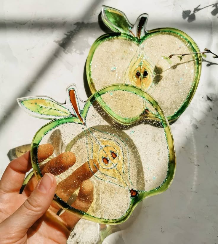 9f0eda07-ddec-48f7-a130-8f668148dd1a Two translucent apple-shaped glass coasters held in sunlight, with intricate seed and leaf details. | Sky Rye Design Two translucent apple-shaped glass coasters held in sunlight, with intricate seed and leaf details.
