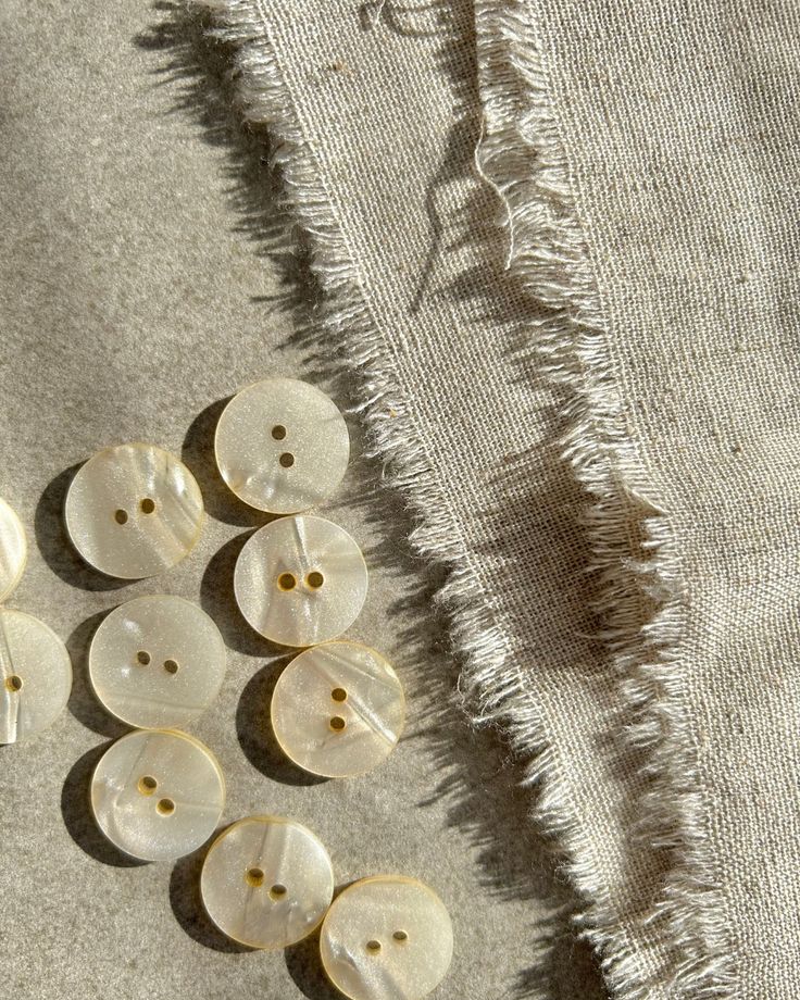 A delicadeza dos aviamentos escolhidos ✨__Conheça em breve todos os detalhes da coleção___09_07 marque esta data 🌊 Cream buttons on frayed beige fabric, sunlight casting gentle shadows, perfect for sewing and crafts. | Sky Rye Design Cream buttons on frayed beige fabric, sunlight casting gentle shadows, perfect for sewing and crafts.