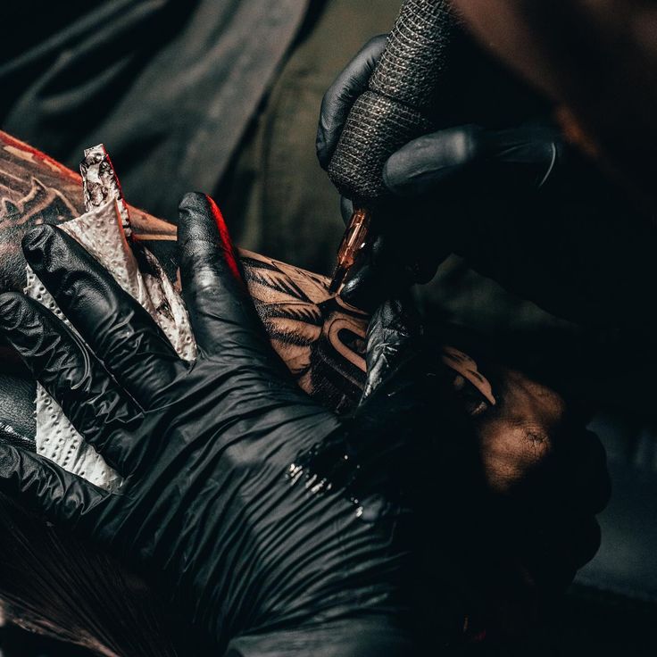Tattoo artist wearing black gloves creates intricate design on skin using a tattoo machine.