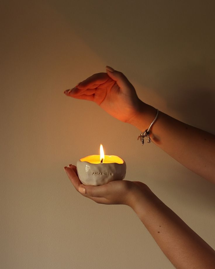Hands holding a lit candle in a ceramic holder against a neutral background.