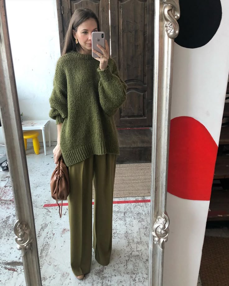 Woman in oversized olive sweater and pants takes mirror selfie, holding a brown bag. Fashionable casual outfit.