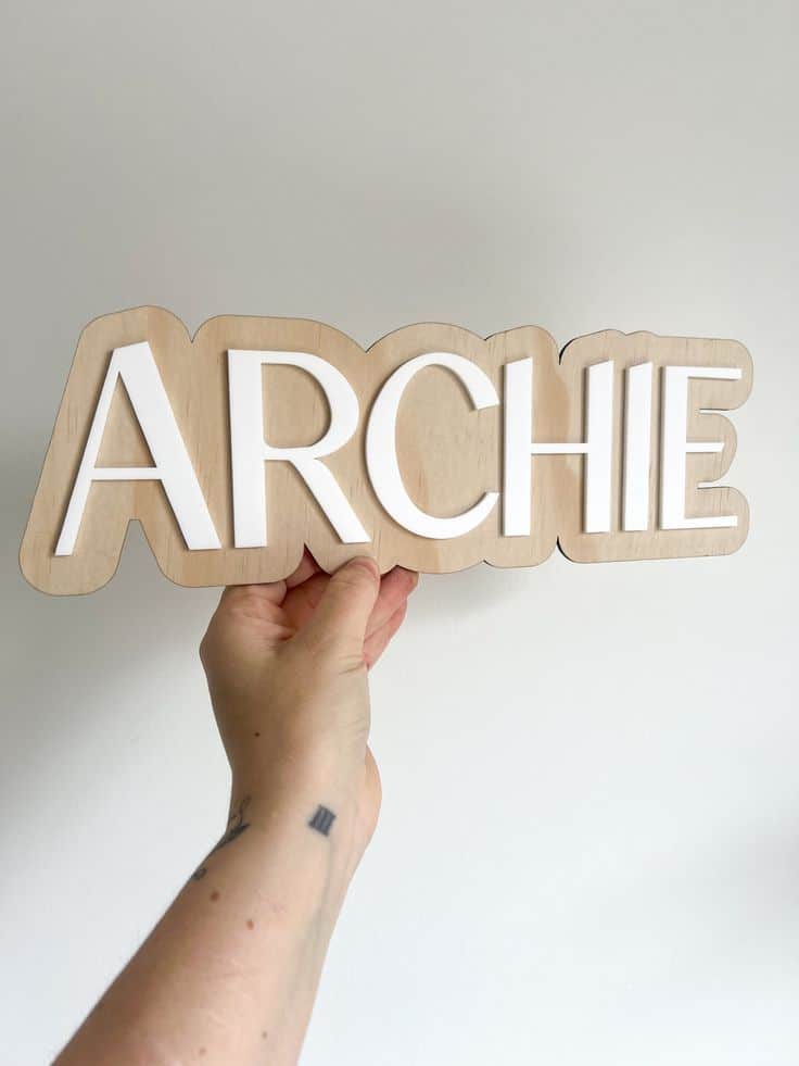 Person holding a wooden sign with the name 'Archie' in white letters against a plain background.