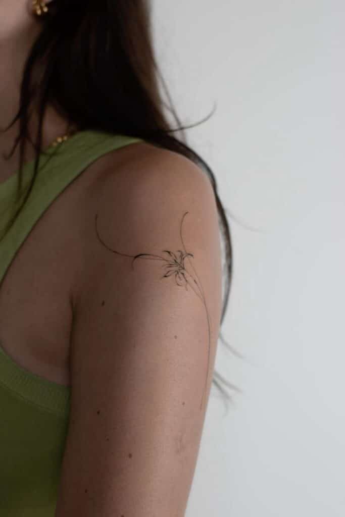 Minimalist floral tattoo on upper arm of person wearing a green top, showcasing delicate lines and curves.