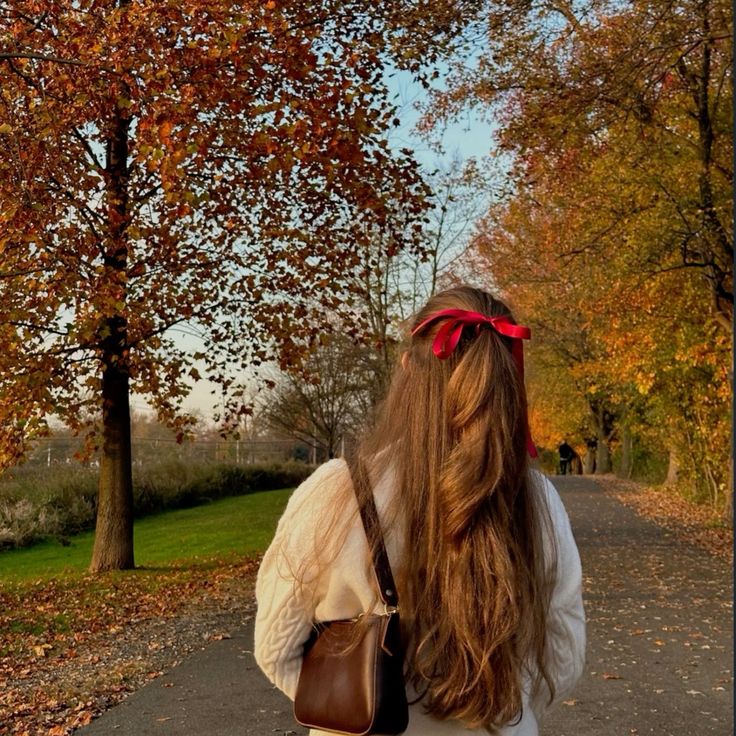 Woman with long hair and red bow walks on autumn path, surrounded by colorful trees.