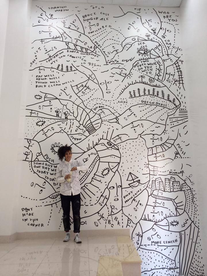 Person standing in front of a large abstract mural with doodles and text on a white wall.