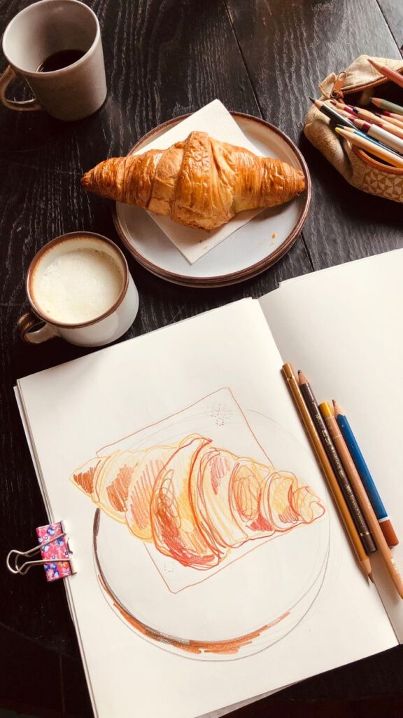 Breakfast scene: croissant with coffee, and sketchbook drawing on table.