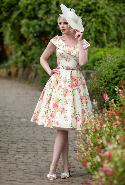 Elegant woman in floral dress and hat poses on garden path with vibrant pink flowers.
