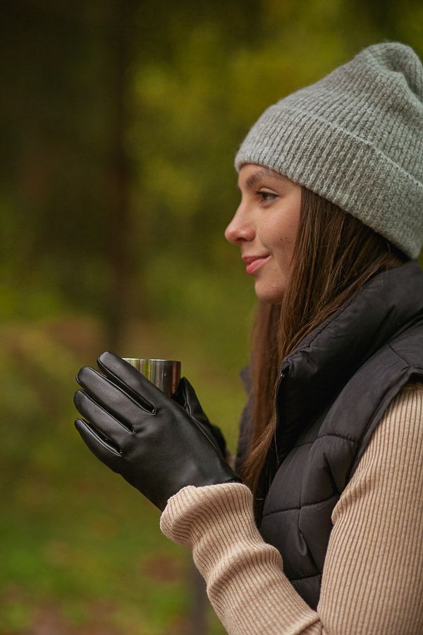 Woman wearing gray beanie, black gloves, and vest holds a cup in a forest setting.