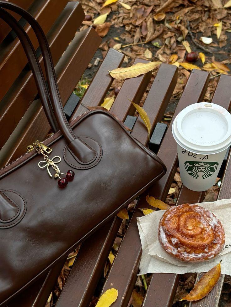 Brown leather handbag, coffee cup, and pastry on a park bench with fallen autumn leaves. Cozy fall setting.