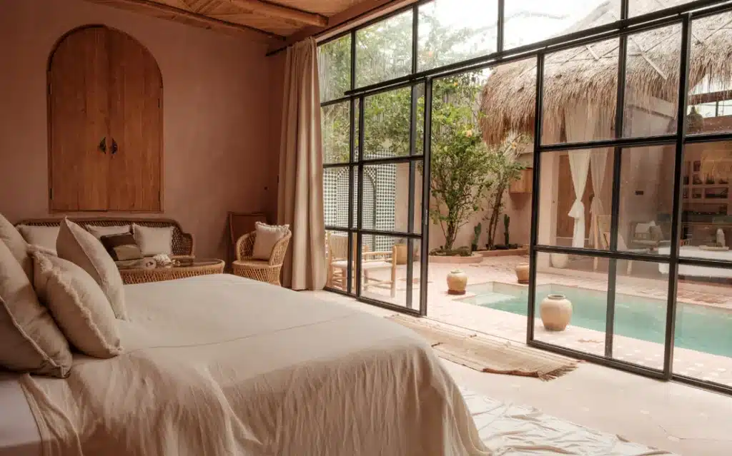Luxury bedroom with view of patio and pool through large windows, featuring natural tones and cozy rattan furniture.