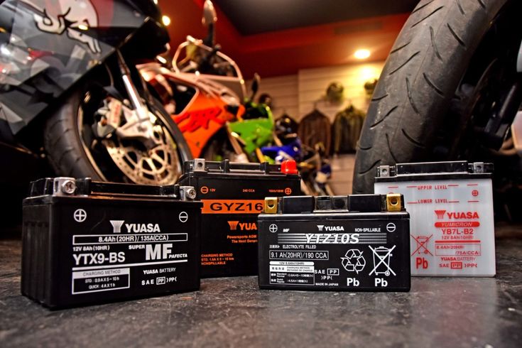Motorcycle batteries displayed in a showroom with bikes in the background, highlighting various Yuasa models.