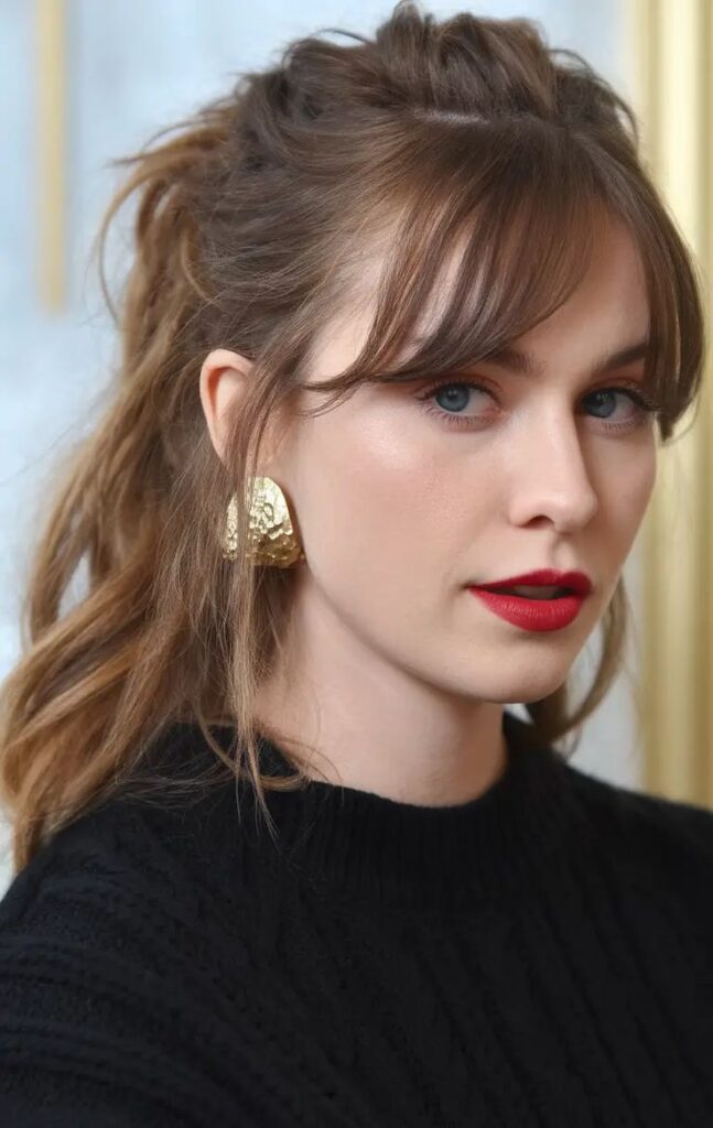 Beautiful Bangs Hairstyles for Summer 2025 Curtain Wispy Curly Easy Fringe Looks Woman with red lipstick, gold earrings, wavy brown hair in ponytail, black sweater, neutral background. | Sky Rye Design Woman with red lipstick, gold earrings, wavy brown hair in ponytail, black sweater, neutral background.