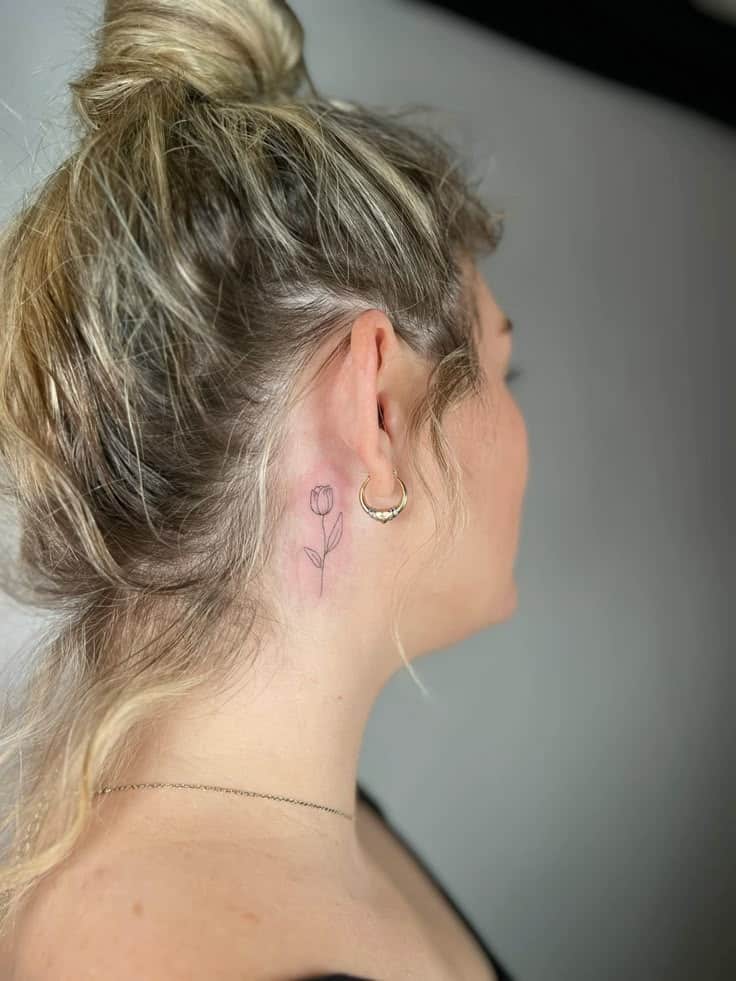 Blonde woman with small flower tattoo behind ear, gold hoop earring, hair in a bun.