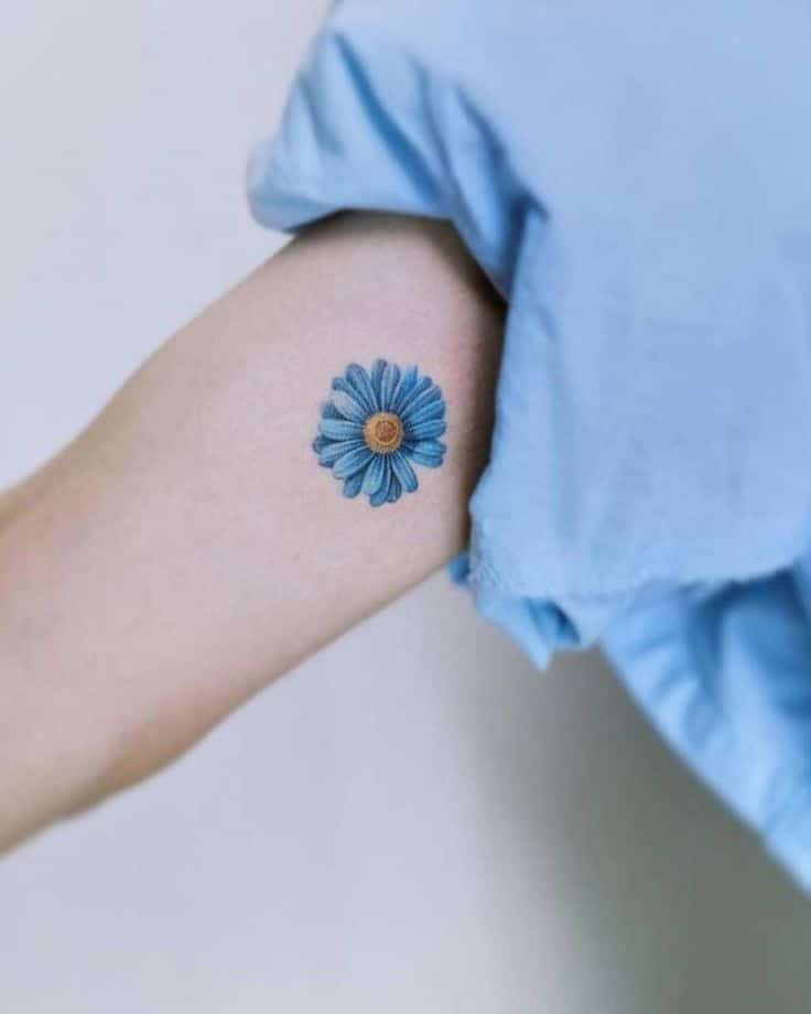 Arm with blue daisy tattoo and blue shirt, minimal floral design on skin.