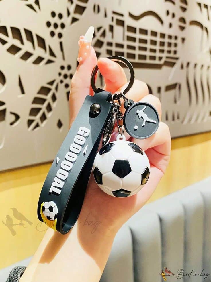 Hand holding a football-themed keychain with a mini soccer ball and GoaaaL strap, against patterned background.