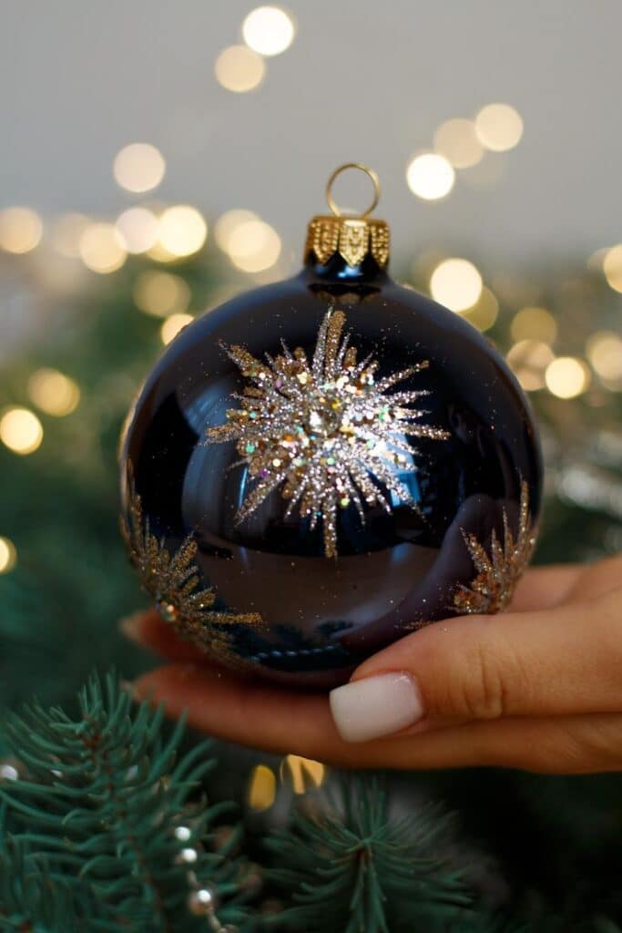 Elegant blue Christmas ornament with gold glitter design held by hand, against a blurred festive light backdrop.