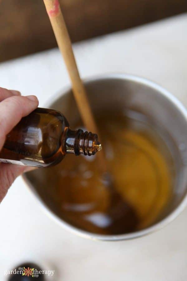 Pouring essential oil from brown bottle into mixture in a metal pot with wooden stick stirrer for homemade recipe.