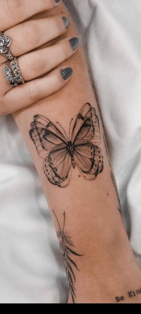 Delicate butterfly tattoo on forearm, accompanied by stylish rings and dark nail polish.
