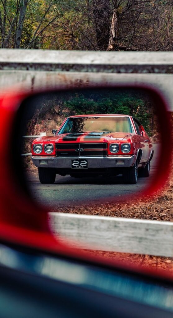 Red classic car viewed through side mirror on a scenic road surrounded by trees.