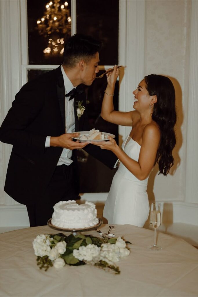 Bride and groom laughing while feeding each other cake at wedding reception.