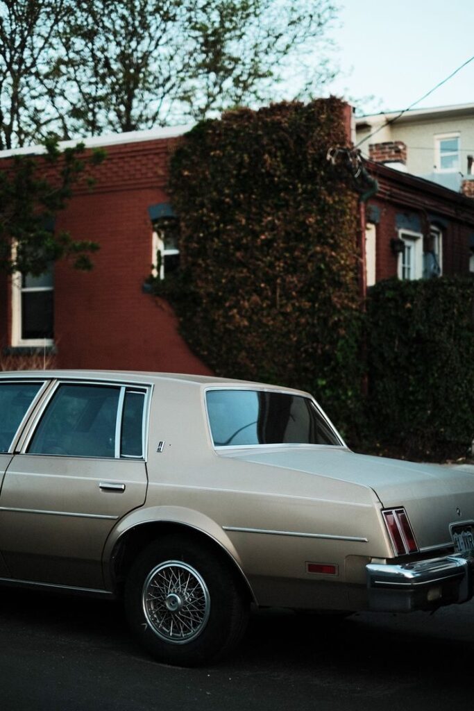 Vintage beige car parked beside ivy-covered brick house.