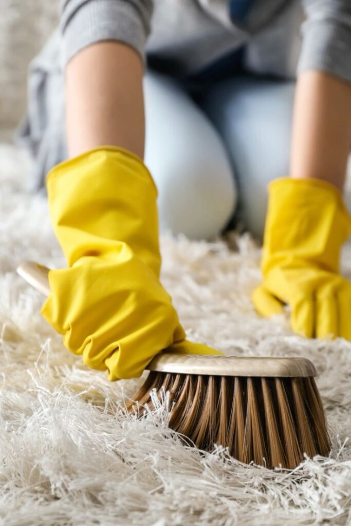 Person scrubbing a carpet with a brush while wearing yellow rubber gloves for deep cleaning.