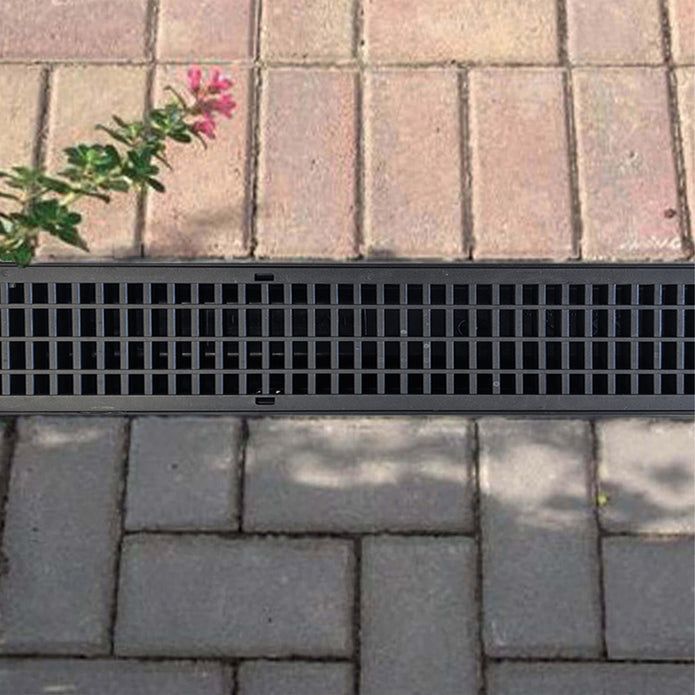 Black metal drainage grate installed on a brick patio with a pink flower visible, casting a shadow on the pavement.