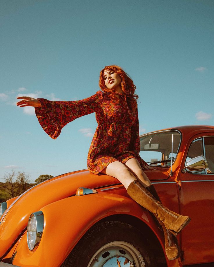 Woman in a floral dress sits on orange vintage car under clear blue sky, expressing carefree joy.