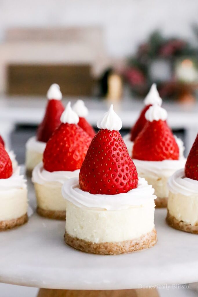 Mini cheesecakes topped with strawberry Santas, perfect for festive holiday desserts on a white serving tray.