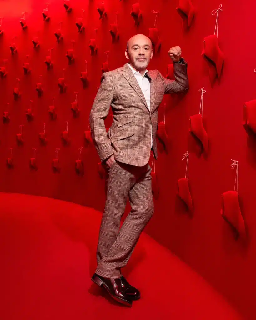 Christian-Louboutin-in-the-Exhibitionniste-exhibition-©-Courtesy-of-Christian-Louboutin Man in plaid suit posing against a red wall with hanging art pieces. | Sky Rye Design Man in plaid suit posing against a red wall with hanging art pieces.