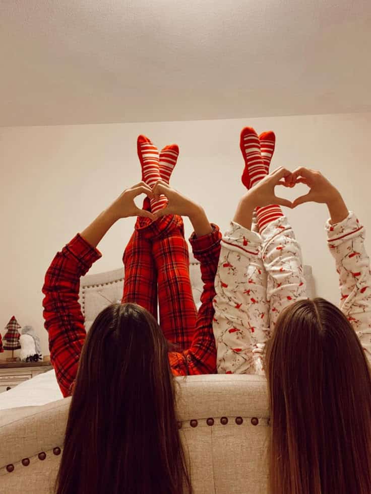 Two people in festive pajamas make heart shapes with hands, feet up, lying on a bed, expressing holiday spirit and fun.