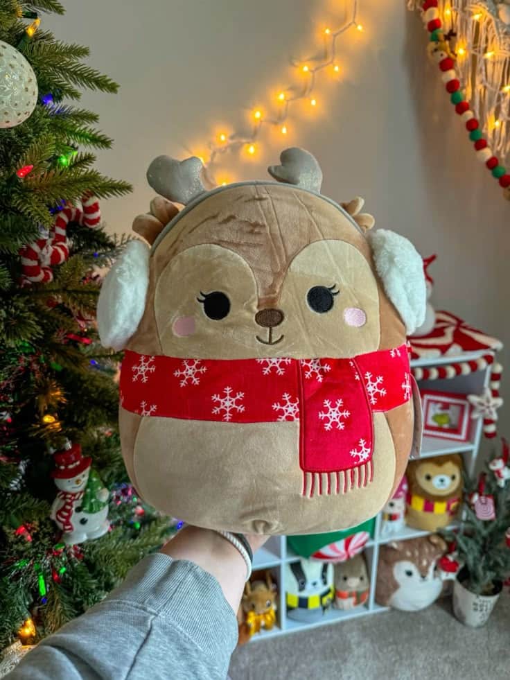 Cute reindeer plush toy with scarf, holiday decorations, and Christmas tree in cozy room.