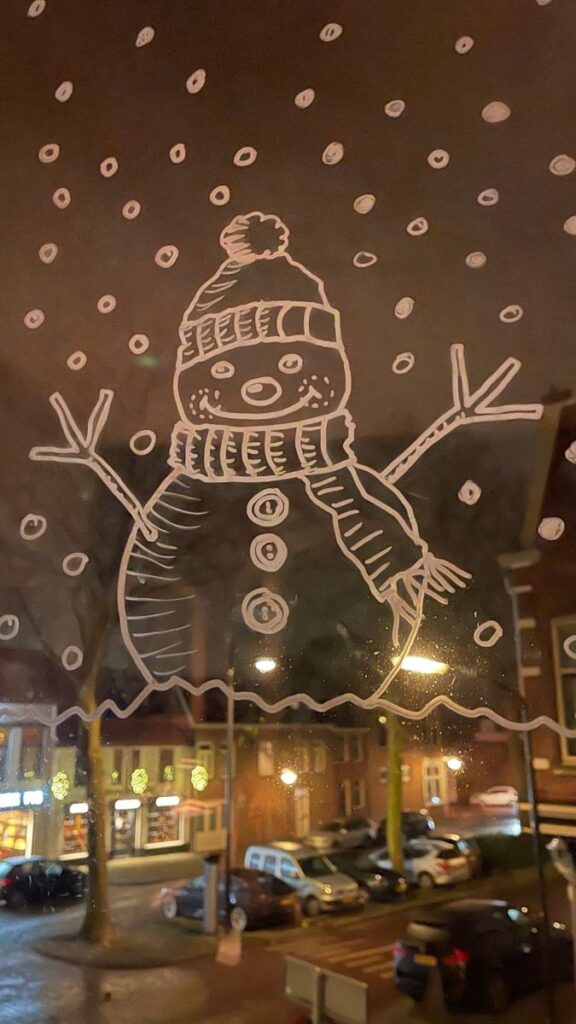 Snowman drawing on a window with festive street view at night.
