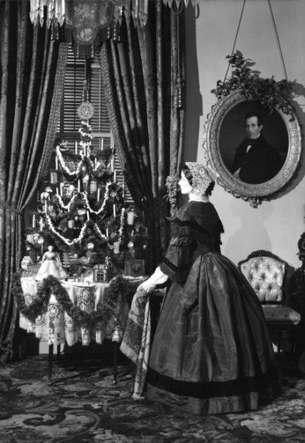 Victorian woman in period dress gazes at an ornate Christmas tree in an elegant parlor with vintage decor.