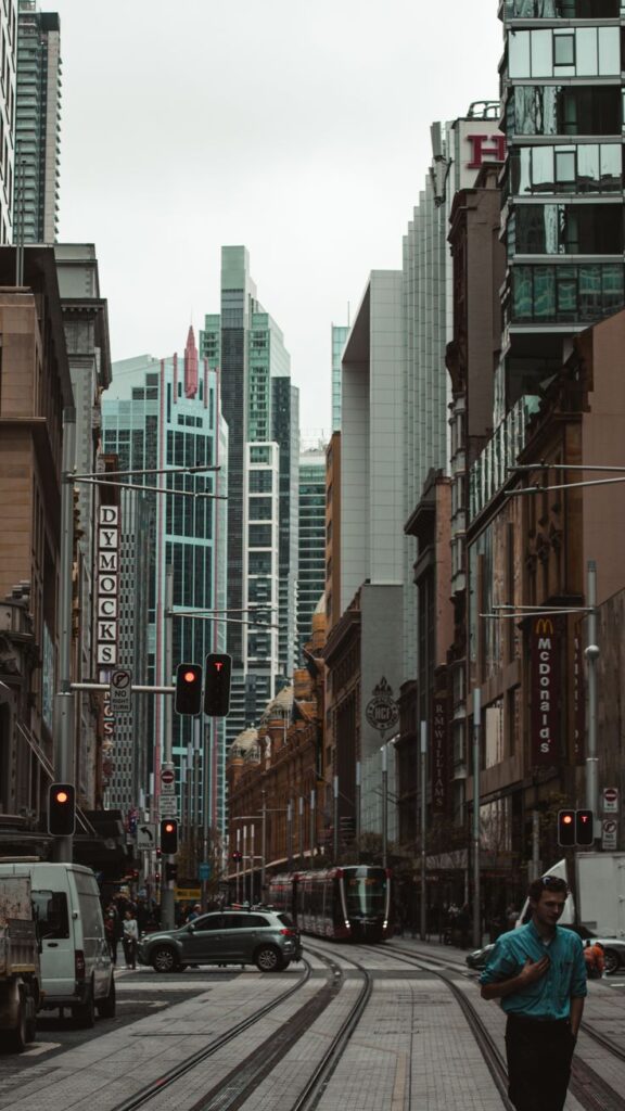 Bustling city street with modern skyscrapers, cars, and pedestrians under an overcast sky. Urban scene with tram tracks.
