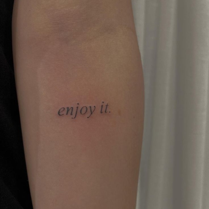 Minimalist arm tattoo reading enjoy it in small black letters.