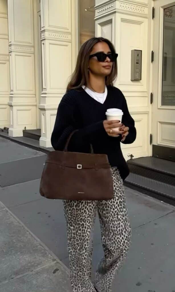 Stylish woman in sunglasses and leopard pants holding a coffee and brown bag on a city street.