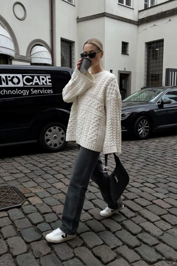 Fashionable woman in cozy sweater and jeans walks with coffee on cobblestone street.