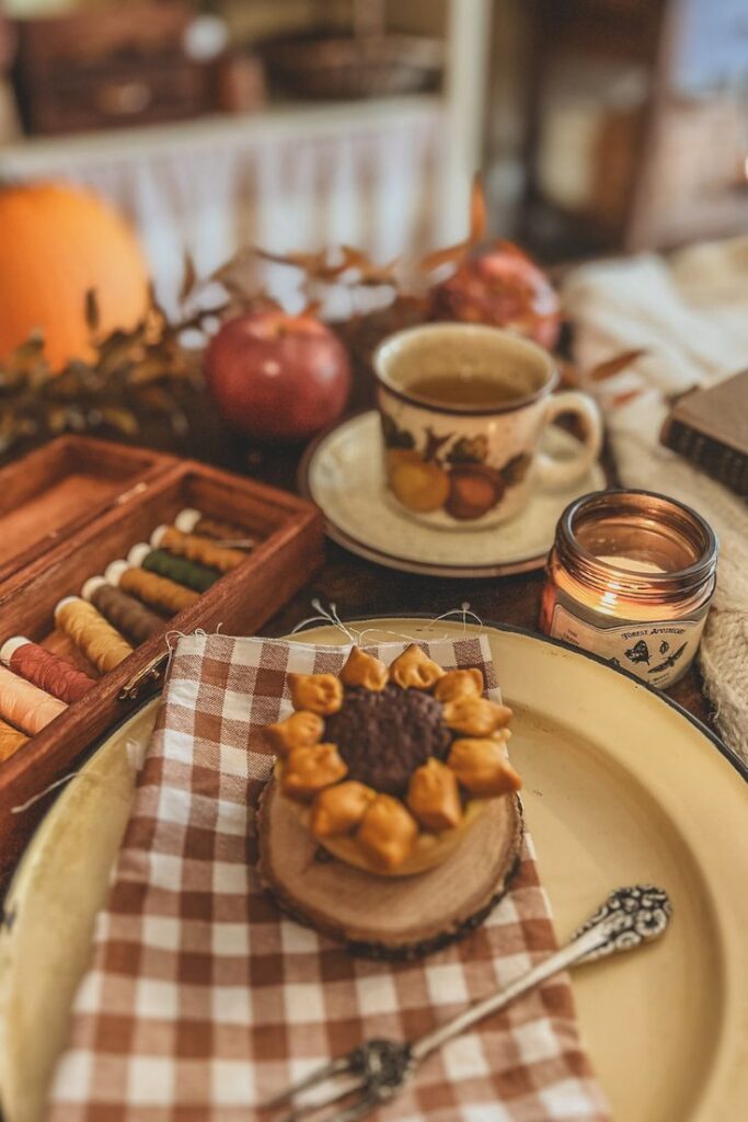 Cozy autumn setup with tea, a candle, sewing threads, and sunflower-themed decor on a checkered napkin.