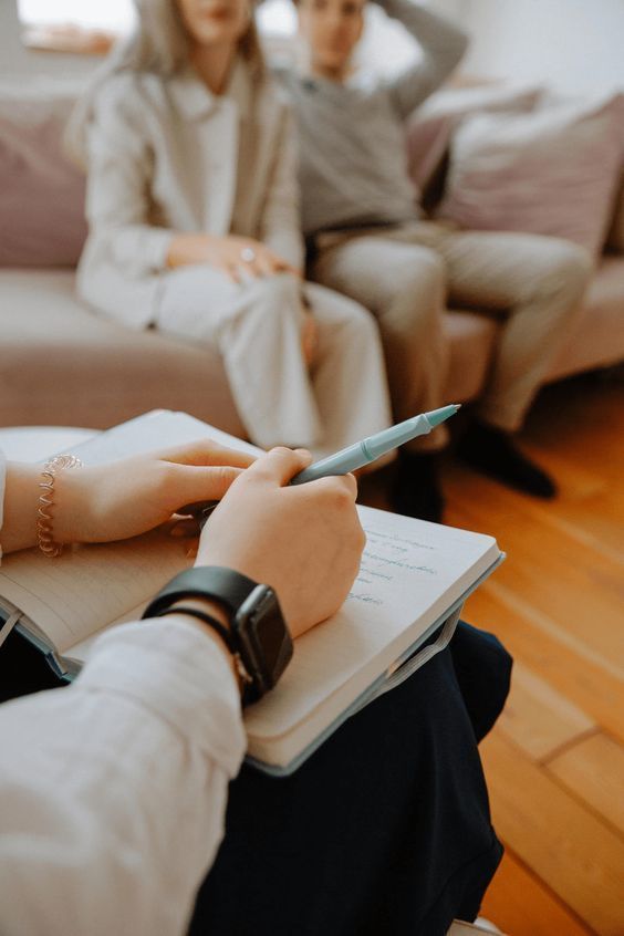 Counselor taking notes during a therapy session with a couple on a sofa nearby.