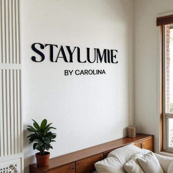 Modern living room with Staylumie by Carolina sign, featuring cozy sofa, wooden cabinet, plant, and window view.
