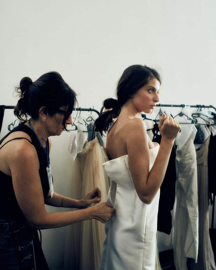 Designer adjusts elegant white dress on model backstage at fashion show.