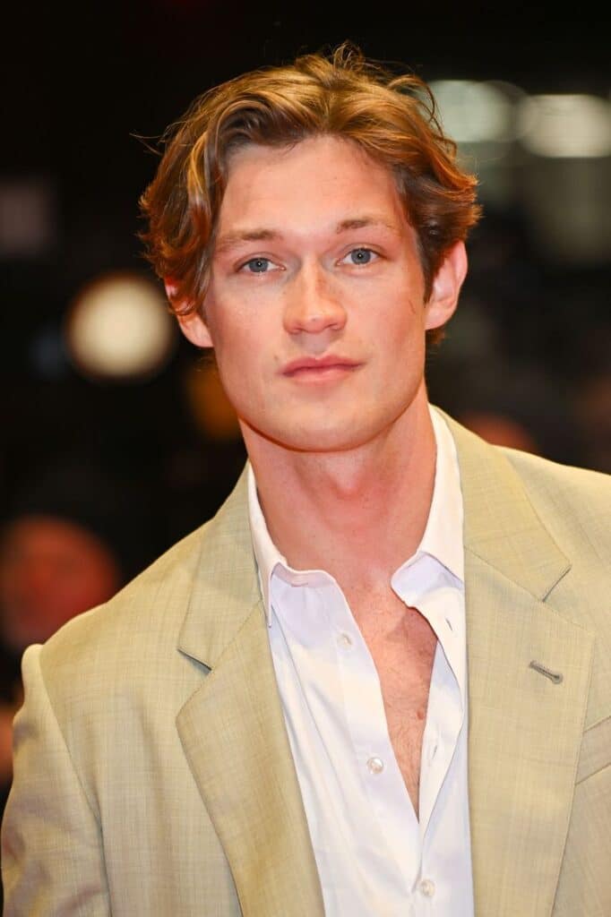 Young man in a beige suit and white shirt, standing on a red carpet with a blurry background.